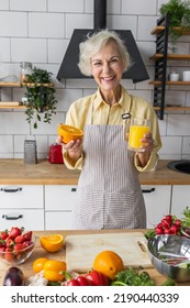 Attractive Senior Woman Drinking Freshly Squeezed Orange Juice, Looking Happy And Healthy. Concept Of Active Lifestyle Of Mature People, Cooking Organic Food. Mature Female At Home At Her Kitchen