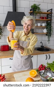 Attractive Senior Woman Drinking Freshly Squeezed Orange Juice, Looking Happy And Healthy. Concept Of Active Lifestyle Of Mature People, Cooking Organic Food. Mature Female At Home At Her Kitchen