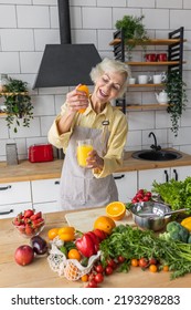 Attractive Senior Woman Drinking Freshly Squeezed Orange Juice, Looking Happy And Healthy. Concept Of Active Lifestyle Of Mature People, Cooking Organic Food. Mature Female At Home At Her Kitchen