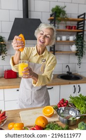 Attractive Senior Woman Drinking Freshly Squeezed Orange Juice, Looking Happy And Healthy. Concept Of Active Lifestyle Of Mature People, Cooking Organic Food. Mature Female At Home At Her Kitchen