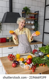 Attractive Senior Woman Drinking Freshly Squeezed Orange Juice, Looking Happy And Healthy. Concept Of Active Lifestyle Of Mature People, Cooking Organic Food. Mature Female At Home At Her Kitchen