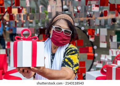 Attractive Senior Woman With Gift In Hand. Portrait An Older Woman With A Gift Box. Elderly Woman Wear Glasses And Fabric Mask Holding A Gift Box