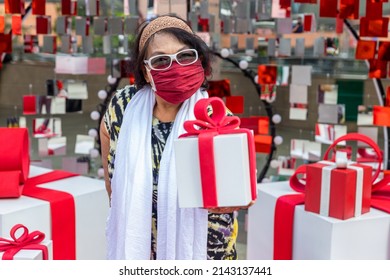 Attractive Senior Woman With Gift In Hand. Portrait An Older Woman With A Gift Box. Elderly Woman Wear Glasses And Fabric Mask Holding A Gift Box