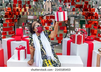 Attractive Senior Woman With Gift In Hand. Portrait An Older Woman With A Gift Box. Elderly Woman Wear Glasses And Fabric Mask Holding A Gift Box