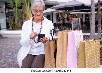 Attractive senior woman relaxing after shopping using mobile phone. Smiling elderly woman with eyeglasses and necklace sitting close to their shopping bags.  Consumerism, shopping, lifestyle concept