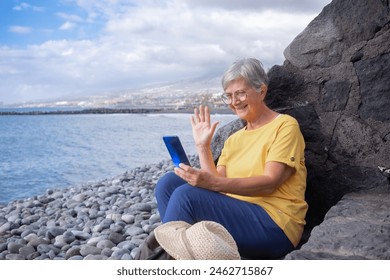 Attractive senior woman sitting on a pebble beach at sea using mobile phone in video call. Mature woman smiles holding cellphone waving hand. Concept of vacation, freedom and happy retirement - Powered by Shutterstock