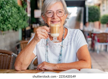 Attractive senior woman sitting outdoor at a cafe table enjoying a coffee and milk drink - caucasian elderly lady with glasses relaxed in retirement or vacation - Shutterstock ID 2231420149
