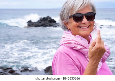 Attractive Senior Woman With White Hair Removes Her Face Mask Against Covid-19 Infection And Enjoys Freedom By The Sea. Active And Smiling Retired