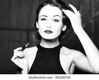 Attractive sensual fashionable retro elegant young adult woman with classic hairstyle and red lips holding lipstick in evening dress indoor on wooden background, horizontal picture