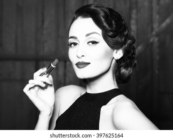 Attractive sensual fashionable retro elegant young adult woman with classic hairstyle and red lips holding lipstick in evening dress indoor on wooden background, horizontal picture