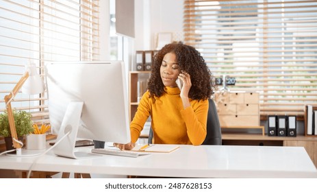 An attractive, serious Black businesswoman is working on the computer while talking on the phone with her business client at her desk in the office. dealing business over the phone, answering the call - Powered by Shutterstock