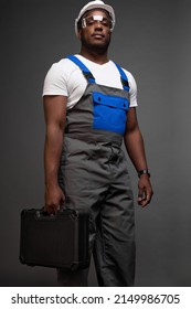 An attractive serious black worker stands in confident posture with suitcase of tools in his hand. Photo from below of tall muscular hard worker in gray jumpsuit with blue pocket and straps