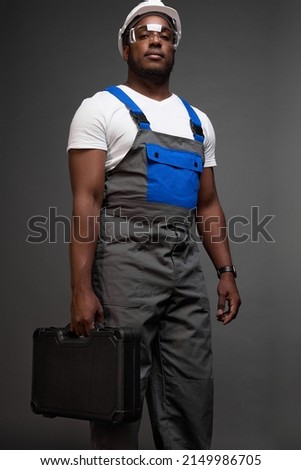 An attractive serious black worker stands in confident posture with suitcase of tools in his hand. Photo from below of tall muscular hard worker in gray jumpsuit with blue pocket and straps