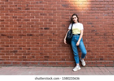 Attractive serious young brunette girl in blue jeans and yellow t-shirt, with black shoulder bag isolated over red brick wall, outdoor photo