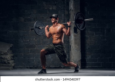 Attractive Shirtless Man In Cap Is Doing Lunge With Barbell At Dark Photo Studio.