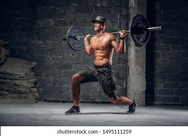 Attractive Shirtless Man In Cap Is Doing Lunge With Barbell At Dark Photo Studio.