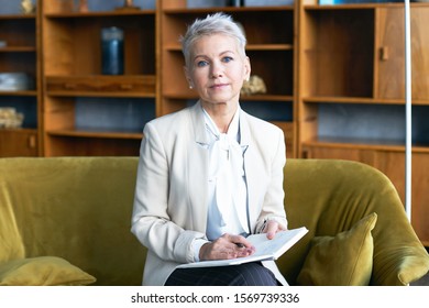 Attractive Short Haired Middle Aged Female CEO Sitting On Comfortable Couch In Office Interior Writing In Her Notebook, Checking Appointment List, Looking At Camera With Serious Confident Expression