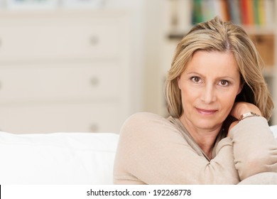 Attractive Sincere Middle-aged Woman Sitting On A Sofa Leaning Her Head On Her Raised Arm Looking Directly At The Camera With A Serious Expression