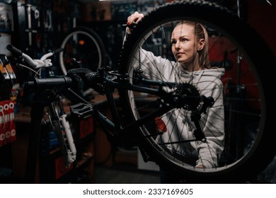 Attractive skilled cycling repairman female repairing and fixing mountain bicycle standing on bike rack in repair workshop with dark interior. Concept of professional maintenance of bicycle transport. - Powered by Shutterstock