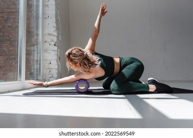 Attractive slim girl with wavy blond hair using foam roller for relaxing muscles on arms after workout. Young fit woman in green sport clothing massaging body on black yoga mat.