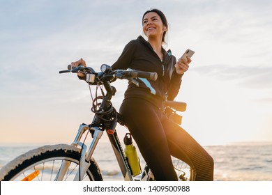 attractive slim woman riding bicycle, sport on morning sunrise beach in fitness suit, healthy lifestyle, listening to music on wireless earphones holding smartphone, relaxing smiling happy - Powered by Shutterstock