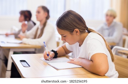 Attractive Smart Teen Girl Studying In Classroom, Listening To Lecturer And Writing In Notebook