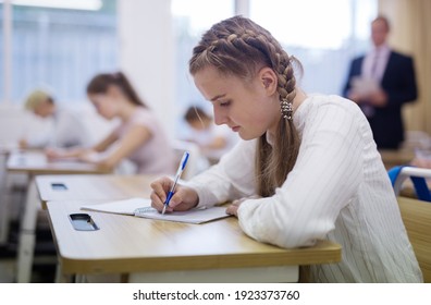 Attractive Smart Teen Girl Studying In Classroom, Listening To Lecturer And Writing In Notebook