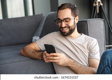 Attractive smart young man sitting on a floor in the living room, using mobile phone