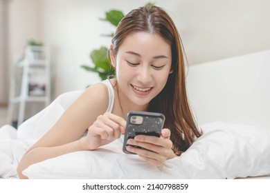 Attractive Smile Pretty Asian Young Woman Using And Playing Mobile Phone, Smartphone Have Video Call Or Chat, Lying Down Relax In Bedroom Wake Up With Covered Blanket In Cozy White Bed In The Morning