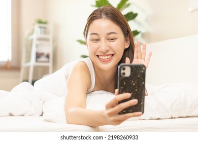 Attractive Smile Pretty Asian Young Woman Using And Playing Mobile Phone, Smartphone Have Video Call Or Chat, Lying Down Relax In Bedroom Wake Up With Covered Blanket In Cozy White Bed In The Morning