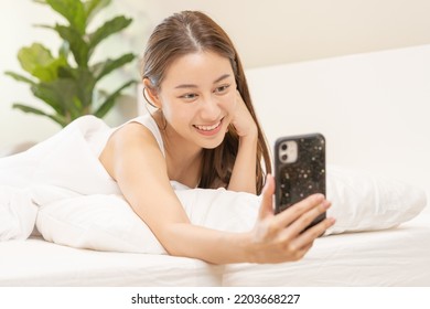 Attractive Smile Pretty Asian Young Woman Using And Playing Mobile Phone, Smartphone Have Video Call Or Chat, Lying Down Relax In Bedroom Wake Up With Covered Blanket In Cozy White Bed In The Morning