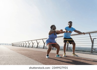 Attractive smiling african american man and woman working out together, squatting with fitness bands outdoors, copy space. Personal trainer training plus size female. Healthy lifestyle concept - Powered by Shutterstock