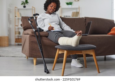 Attractive smiling african american woman works at home in the living room with laptop and makes notes in notebook, sitting on the sofa with broken bandaged leg lying on stool. Crutches are nearby. - Powered by Shutterstock