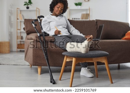 Attractive smiling african american woman works at home in the living room with laptop and makes notes in notebook, sitting on the sofa with broken bandaged leg lying on stool. Crutches are nearby.
