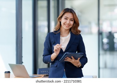 Attractive smiling Asian businesswoman standing holding tablet working and recording work details in office.