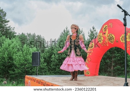 Attractive smiling Asian girl in fur headdress and traditional festive Bashkir clothes performing, dancing on stage. Village festival, Siberia, Russia