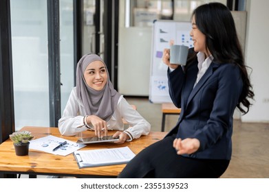 An attractive and smiling Asian Muslim female office worker wearing a hijab enjoys chatting with her colleague during a coffee break in the office. - Powered by Shutterstock