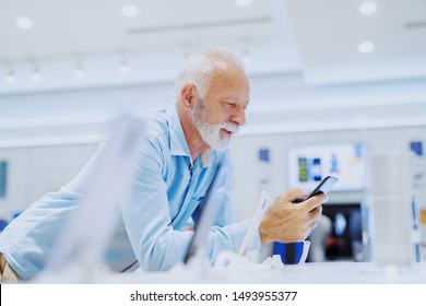 Attractive Smiling Bearded Caucasian Senior Man Leaning On Stand And Trying Out New Smart Phone. Tech Store Interior.