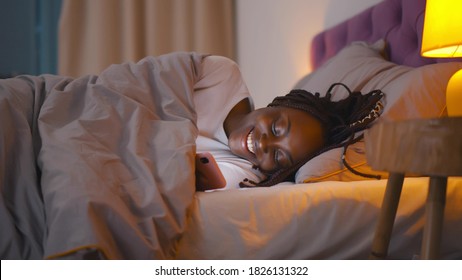 Attractive smiling black young woman using mobile phone while lying in bed in evening. Side view of happy african female chatting with friends on smartphone relaxing in bedroom before sleep - Powered by Shutterstock