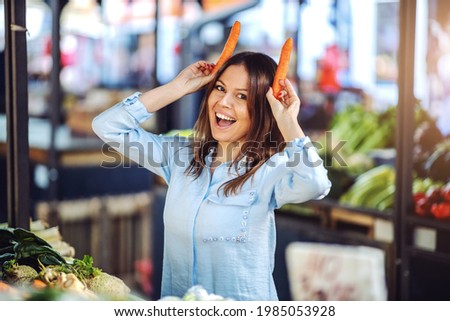 Attractive smiling brunette holding carrots as horns and looking at camera while standing at farmers market.