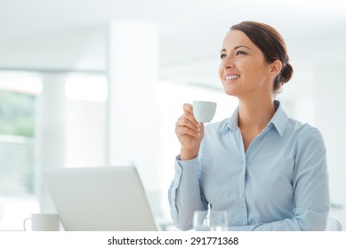 Attractive smiling business woman sitting at office desk, holding a cup of coffee, she is relaxing and looking away