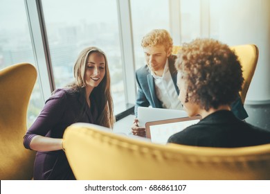 Attractive smiling businesswoman is showing screen of her gadget to her boss and female colleague during work meeting while sitting on yellow armchair in office interior next to window of skyscraper - Powered by Shutterstock