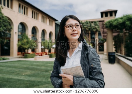 Attractive smiling charming asian young girl student with school book relaxing walking in university outdoor. college lady enjoy sunshine at the park in stanford surrounding by green grass and trees.