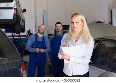 Attractive Smiling Female Insurance Agent And Two Auto Mechanics Near Car At Workshop. Focus On Girl