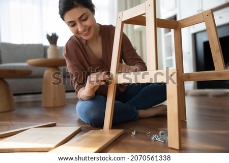 Attractive smiling Indian woman hold screws collects wooden furniture details seated on warm floor in living room, close up, assembling new purchased online wood chair. Interior items store ad concept