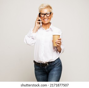 Attractive Smiling Mature Woman With Short Gray Hair Holding Mobile Phone And Cup Of Takeaway Coffee Over Grey Background. Lifestyle, Business And Old People Concept.