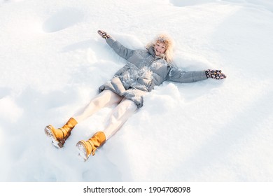 Attractive Smiling Mid Adult Caucasian Woman Wearing Gray Down Jacket And Yellow Boots Lying In Snow Making Snow Angel.