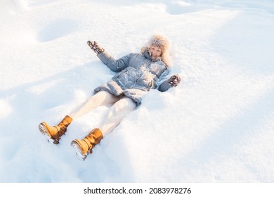 Attractive Smiling Mid Adult Caucasian Woman Wearing Gray Down Jacket And Yellow Boots Lying In Snow Making Snow Angel.