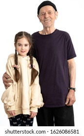 Attractive Smiling Old Man In Black Hat, T-shirt And White Wireless Headphones, Senior Pencioner Looking At The Camera And Hugging His Granddaughter Dressed In Beige Hoodie, White Wall On The Backgrou