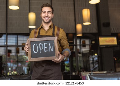 attractive smiling waiter holding chalkboard with open inscription in cafe - Powered by Shutterstock